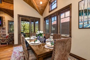 Uma sala de jantar com mesa de madeira posta para seis, janelas amplas e parede de pedra.