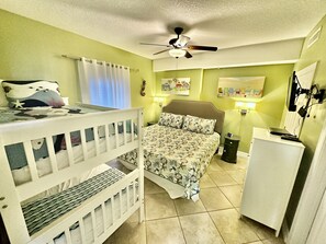 A room with a bunk bed, a single bed with floral bedding, a ceiling fan, and a small white cabinet.