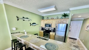 A kitchen with a long island counter, bar stools, a microwave, stove, refrigerator, and decorative fish wall art. - Highly Rated, 2 Bed/2 Bath Ocean Reef Condo, Fabulous Reviews, Beach Service (Panama City Beach)