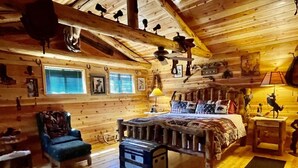 Un dormitorio rústico de cabaña de troncos con una cama, un sillón, una mesita y un tapete de cuero de vaca.