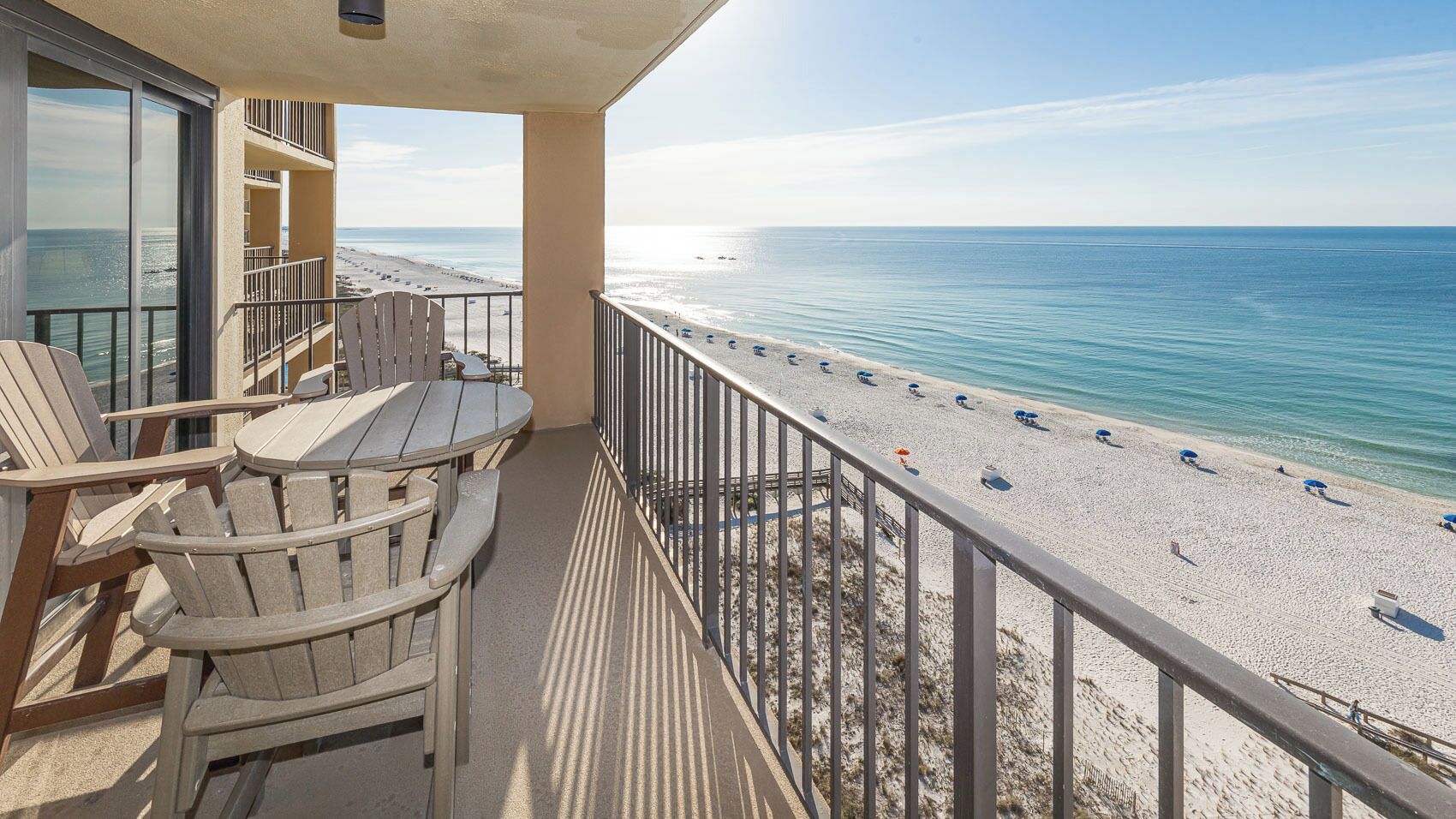 Un balcone con sedie e un tavolo, affacciato su una spiaggia con ombrelloni e vista libera sull'oceano.