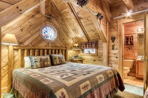 A cozy, wooden cabin bedroom with a large bed, a window with a rose design, and a small table with a lamp. - ER84 - Serenity Summit - Great location. Close to all the action! (Pigeon Forge)