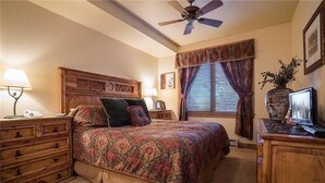 Un dormitorio con cabecera de madera, cama con ropa de cama estampada, mesita de noche con lámpara, escritorio con computadora y ventana con cortinas.