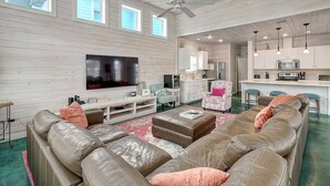 A spacious living room with a large flat-screen TV, leather sofas, a pink armchair, and a kitchen area with modern appliances.