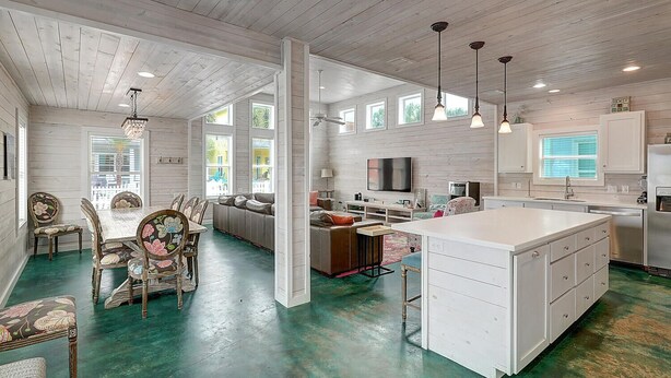 A spacious kitchen with a large island, a dining area with floral chairs, and a living room with a sofa and TV.