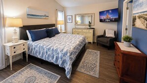 Un dormitorio con cama, mesitas de noche, cómoda, silla, ventilador de techo y obras de arte en las paredes.