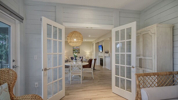A spacious room with a large open door leading to a dining area, a wicker chair, and a bed with a headboard.