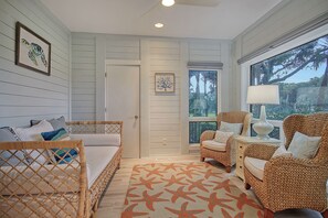 Une chambre avec un lit, deux fauteuils en osier, une table d’appoint avec une lampe, un tapis orné de motifs d’étoiles de mer et une fenêtre donnant sur les arbres.