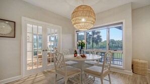 A dining area with a round table, white chairs, a wicker chandelier, and a view through large windows.