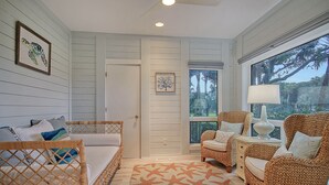 A room with a bed, two wicker chairs, a side table with a lamp, a starfish-patterned rug, and a window with a view of trees.