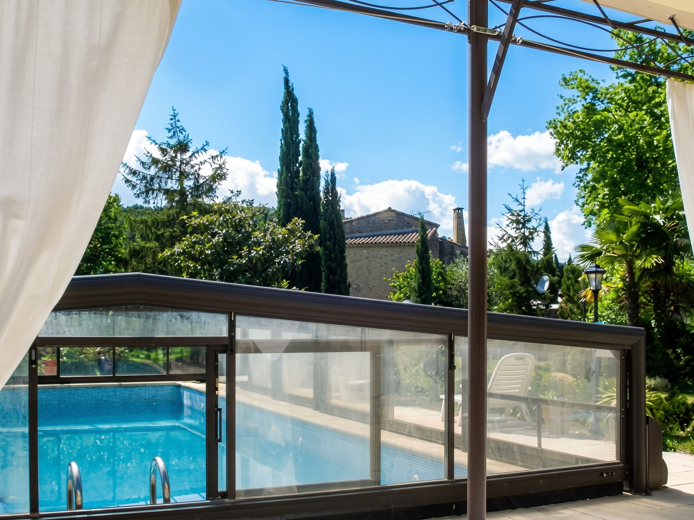 Un espace piscine doté d’une enceinte en verre, offrant une vue dégagée sur la piscine, et un bâtiment avec un toit en tuiles en arrière-plan.