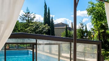 Un espace piscine doté d’une enceinte en verre, offrant une vue dégagée sur la piscine, et un bâtiment avec un toit en tuiles en arrière-plan.