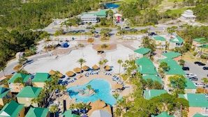 Vista aérea de un complejo turístico con piscina central, varias edificaciones con techos verdes y un paisaje circundante con árboles y una carretera.