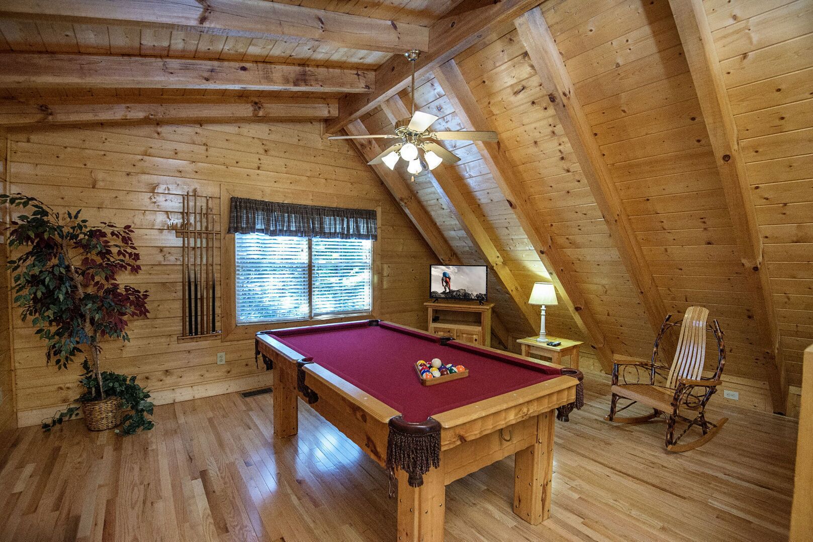 A cozy attic room with a pool table, wooden walls, a ceiling fan, and a window with blinds.