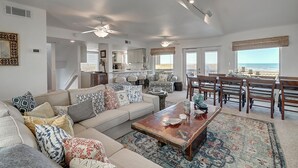 A spacious living room with a large sofa, a coffee table, and a dining area with chairs. There is a view of the ocean through the windows.