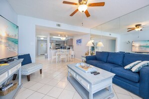 A spacious living room with a blue sofa, a white coffee table, and a TV on a stand. There is a kitchen in the background with white cabinets and a dining area with white chairs.