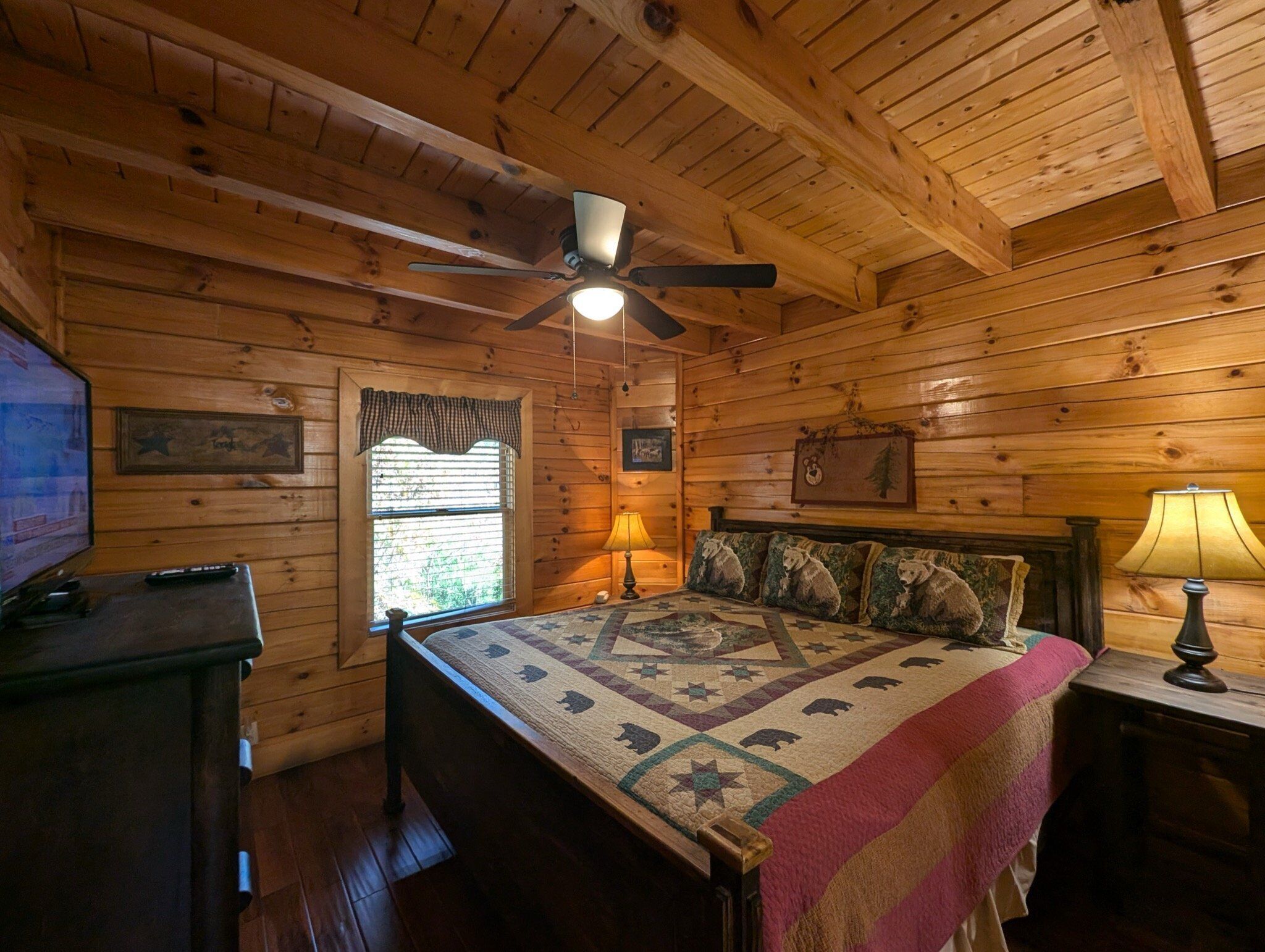 A log cabin bedroom with a bed, a ceiling fan, a window, and a television on a dresser.
