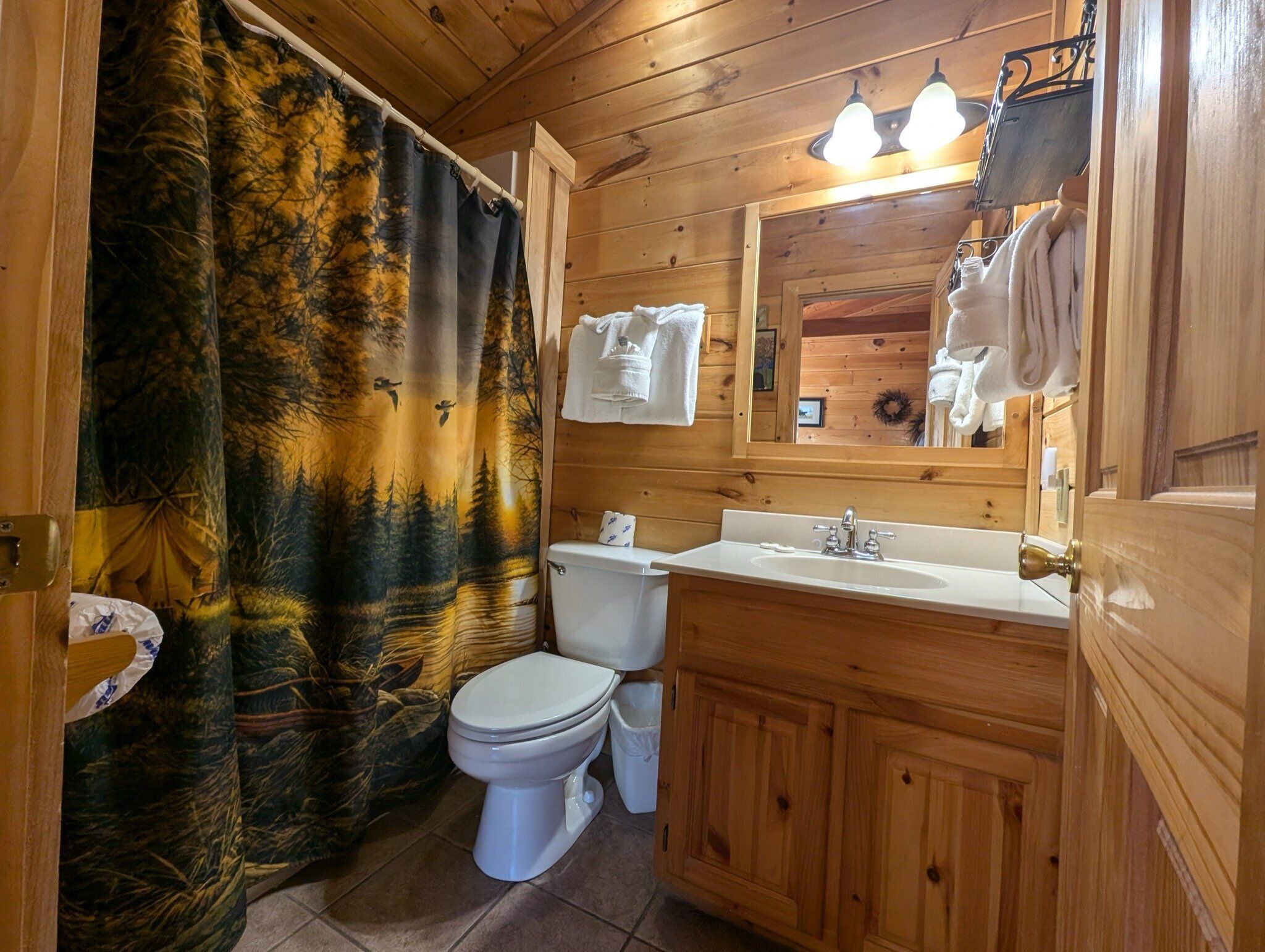 A wooden cabin bathroom with a toilet, sink, and a shower curtain with a forest design.