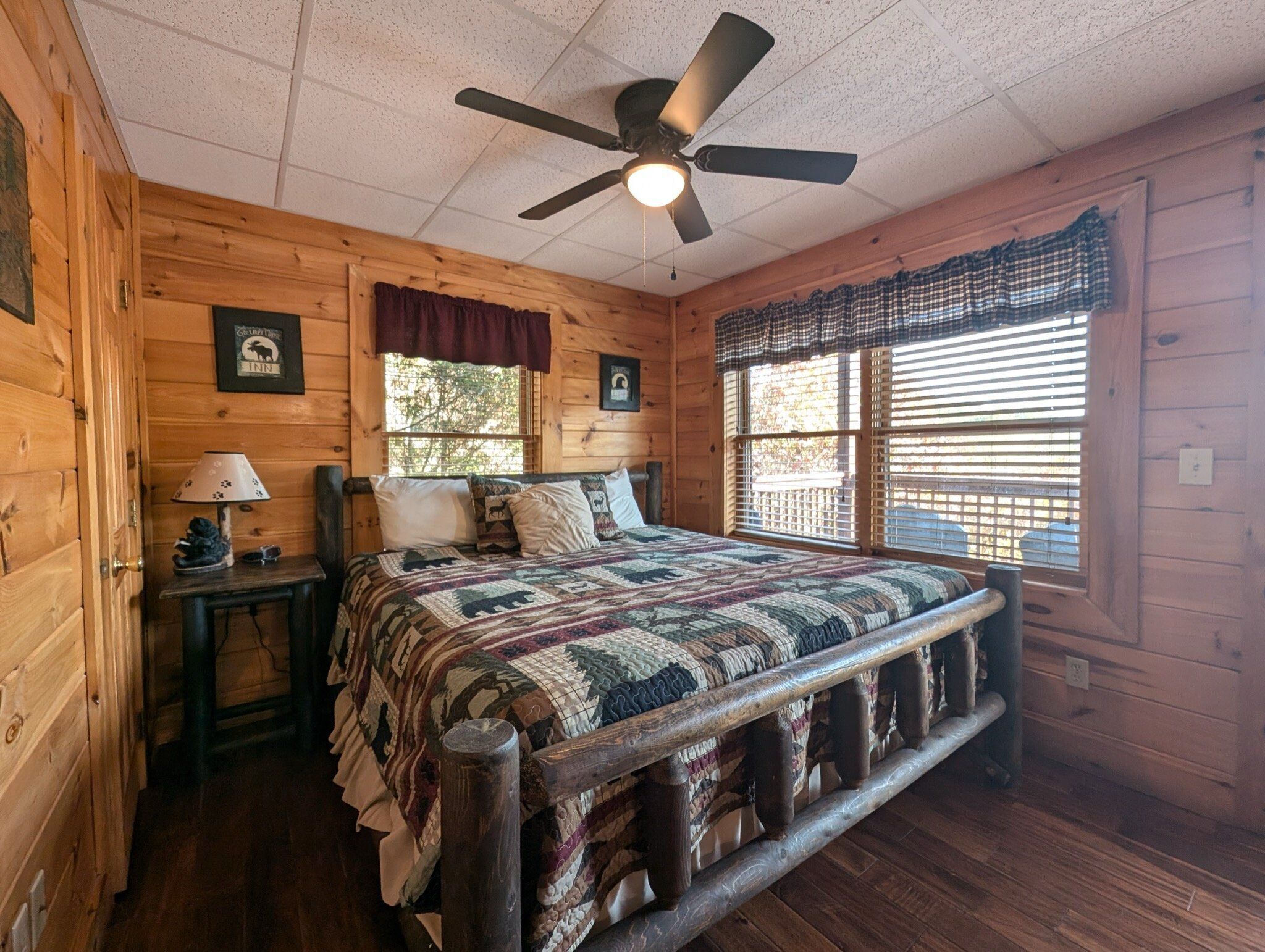 A wooden cabin bedroom with a bed, a nightstand, a lamp, a ceiling fan, and a window with blinds.
