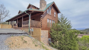 A log cabin with a stone chimney, a porch with a lattice railing, and a view of the surrounding landscape. - ERN807 - Black Bearry Ridge - Great Location! Close To All The Action! (Pigeon Forge)