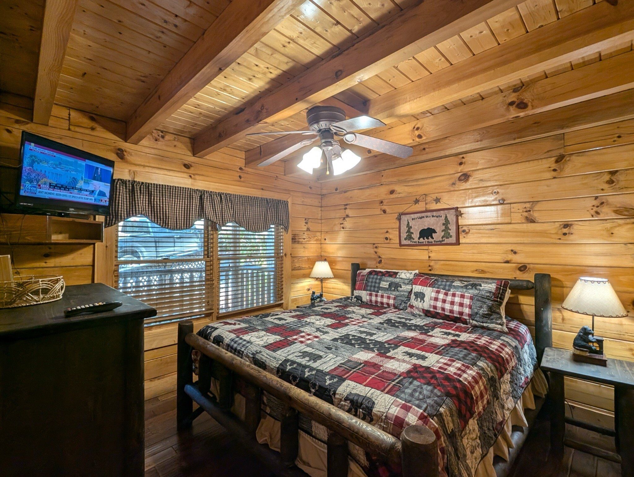 Una habitación cálida tipo cabaña con techo de madera, una cama con colcha, un televisor en un estante y una ventana con persianas.