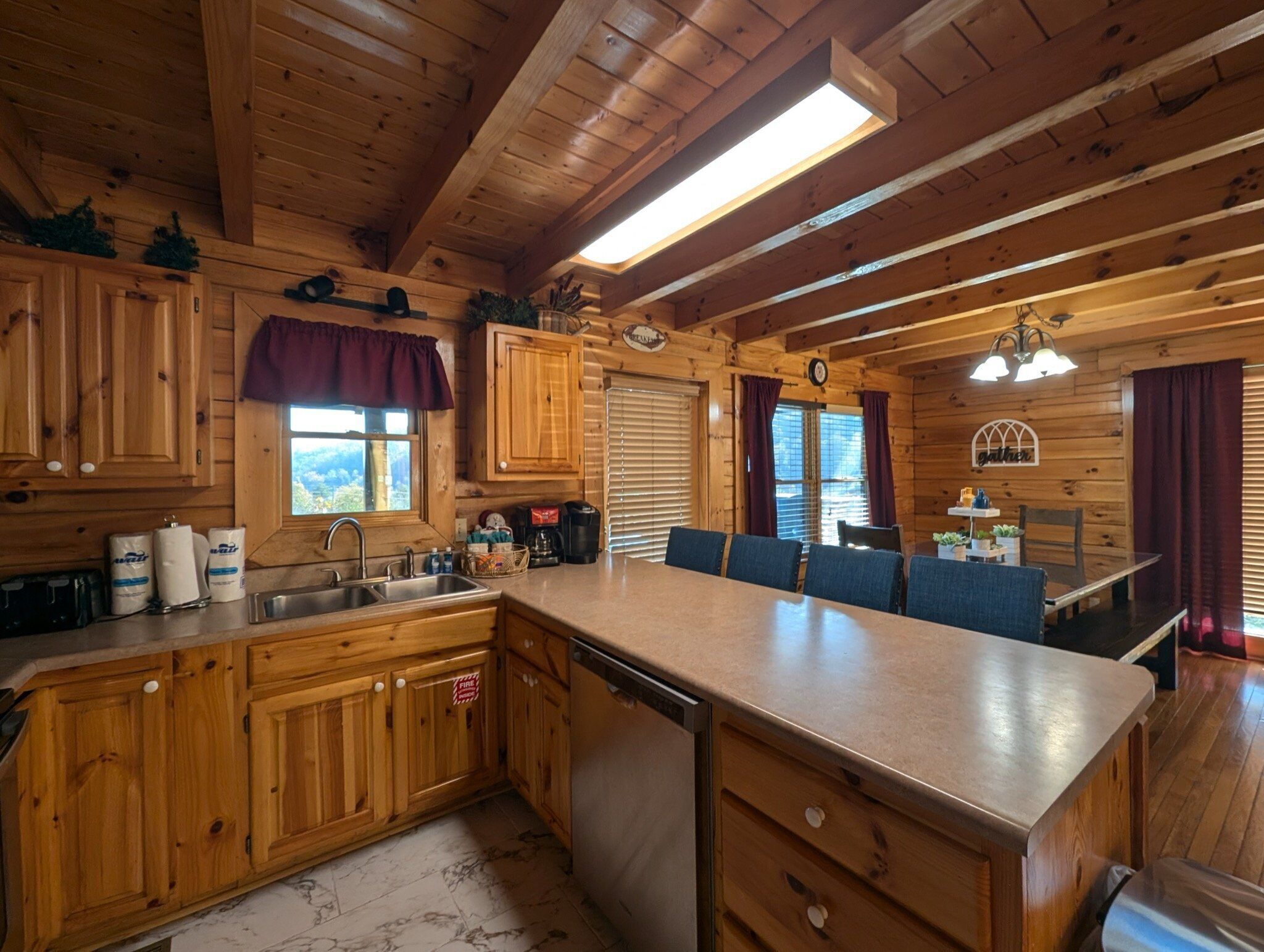 A wooden cabin interior with a kitchen area, dining space, and a large window providing a view of the outdoors.