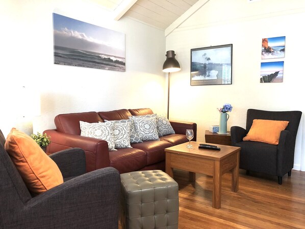 A cozy living room with a leather sofa, a grey armchair, and a wooden coffee table. There are wall-mounted artworks depicting landscapes and a lamp on a side table. - Remodeled Downtown Cottage. Walk to shops, arts, eats and the Carmel Beach (Carmel-by-the-Sea)