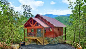 A cabin with a stone foundation, wooden deck, and a metal roof, surrounded by dense forest and mountains.