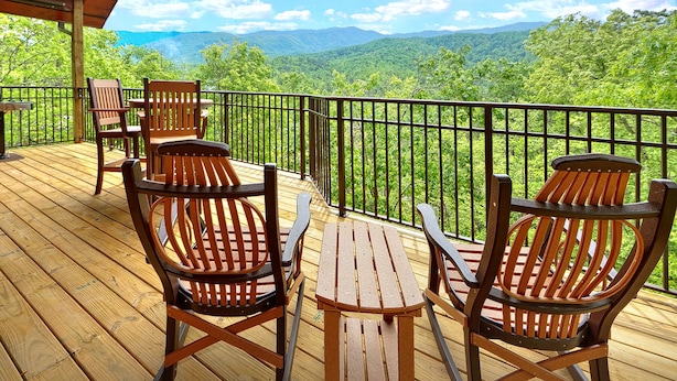 A wooden deck with two Adirondack chairs and a small table, overlooking a lush green landscape with mountains in the distance.