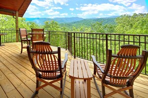 A wooden deck with two Adirondack chairs and a small table, overlooking a lush green landscape with mountains in the distance.