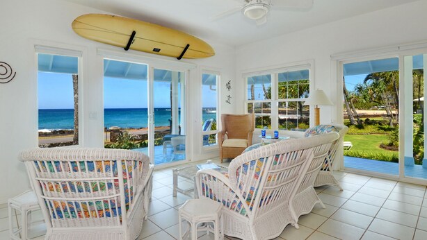 A room with white wicker furniture, a surfboard, and large windows overlooking the beach. - Home Of The Cherished Seacoast By Parrish Kauai- oceanfront & walk to Poipu Beac (Koloa)