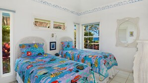 A bedroom with two beds, each with colorful bedding, a large window with a view of the outdoors, and a white wicker chair. - Home Of The Cherished Seacoast By Parrish Kauai- oceanfront & walk to Poipu Beac (Koloa)