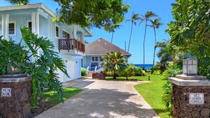 A two-story house with a balcony, a stone wall with a sign, and a well-maintained lawn with palm trees. - Home Of The Cherished Seacoast By Parrish Kauai- oceanfront & walk to Poipu Beac (Koloa)