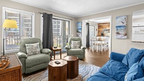 A living room with two green armchairs, a blue sofa, wooden coffee tables, and a dining area with white chairs. - A114 at the Myrtle Beach Resort (Myrtle Beach)