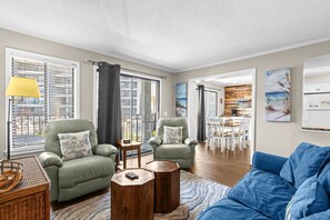 Una sala de estar con dos sillones verdes, un sofá azul, mesas de centro de madera y una zona de comedor con sillas blancas.