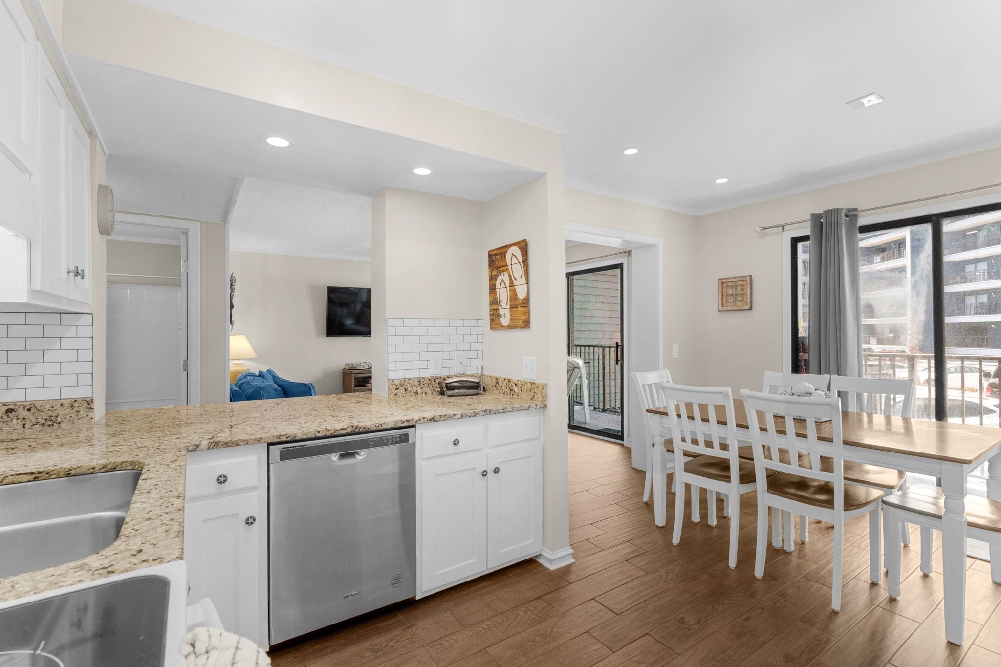 A modern kitchen with white cabinets, granite countertops, and a dishwasher. There is a dining area with white chairs and a wooden table.