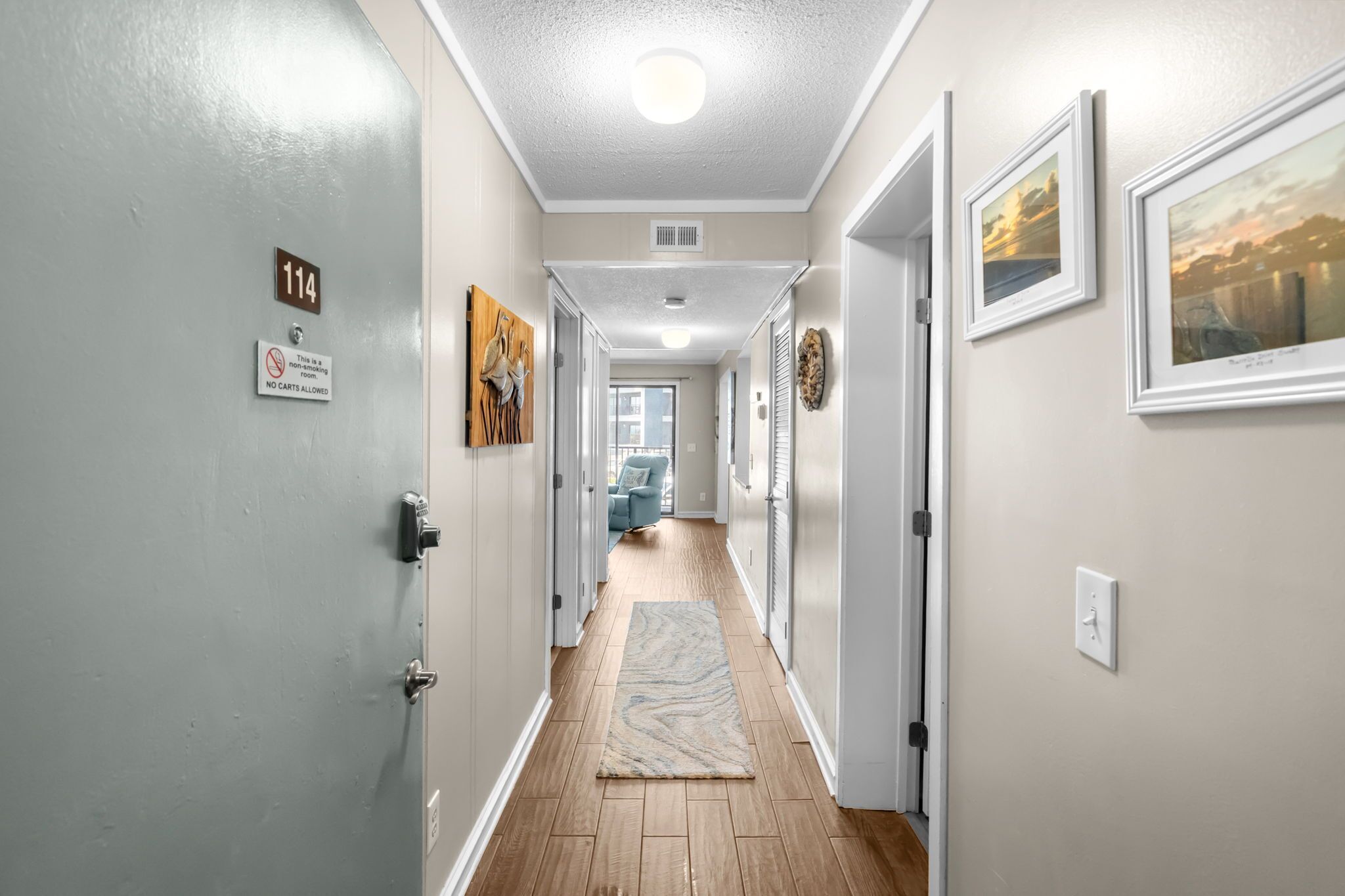A hallway with a wooden floor, a rug, a door with a number sign, and framed pictures on the walls.