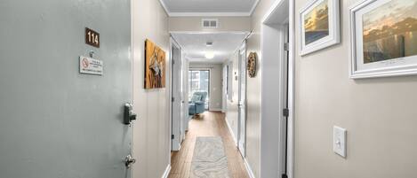 A hallway with a wooden floor, a rug, a door with a number sign, and framed pictures on the walls.