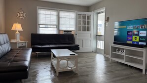 Una sala de estar con un sofá de cuero negro, una mesa de centro blanca, un televisor sobre un mueble bajo y una puerta que da al exterior.