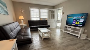 Una sala de estar con un sofá de cuero negro, una mesa de centro blanca, un televisor sobre un mueble, y una puerta que da a un balcón.