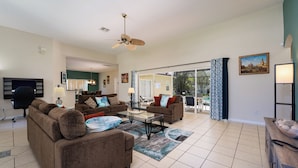 A spacious living room with two sofas, a coffee table, and a ceiling fan. There is a TV mounted on the wall and a window with curtains.