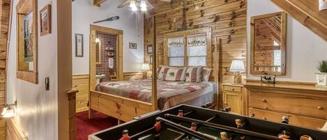 A cozy room with a bed, a foosball table, wooden walls, a ceiling fan, and a window with curtains.