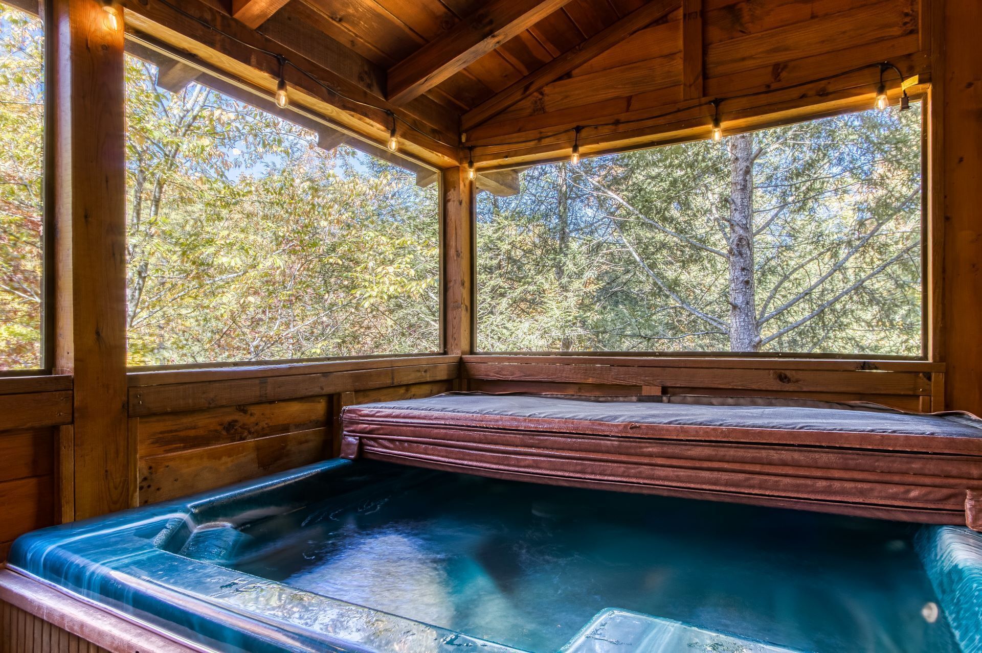 A wooden cabin with a hot tub and large windows overlooking a forest.