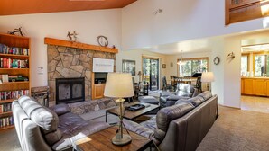 A living room with leather sofas, a stone fireplace, a bookshelf, and a dining area. - BACK03 - Walk to Sunriver Village! Quiet Setting, 2 Suites, A/C, Hot Tub (Sunriver)