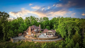 A large, multi-story house surrounded by dense forest, featuring a circular driveway and outdoor seating area. - POOL | Basketball | 6 bdrm | Theater | Acreage! (Gatlinburg)