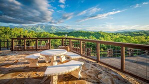 An outdoor patio with stone tables and benches, a metal railing, and a view of green hills and mountains under a partly cloudy sky. - POOL | Basketball | 6 bdrm | Theater | Acreage! (Gatlinburg)