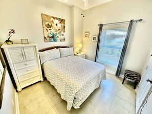 A bedroom with a bed, a dresser, a nightstand, a lamp, a window with blinds, and a painting on the wall.