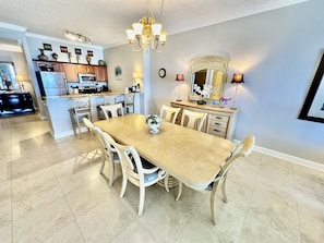 A spacious dining area with a large wooden table and chairs, a chandelier, and a kitchen in the background.