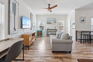 A spacious living room with a wooden floor, a grey sofa, a wooden desk, and a ceiling fan. - Purple Parrot 2nd Floor Condo With Pool (Pensacola)