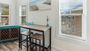 A room with a wooden table and two chairs, a painting of a beach scene, and a view through the window.